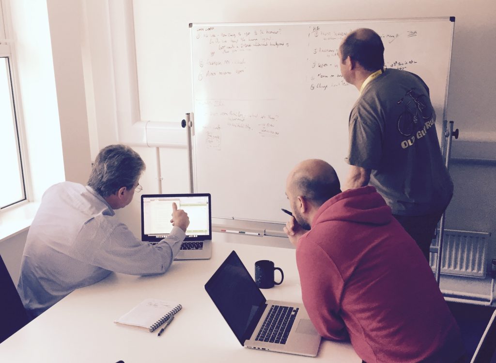 Three people collaborating around laptops and a whiteboard in an office meeting room, discussing software workflows and AI-assisted development.